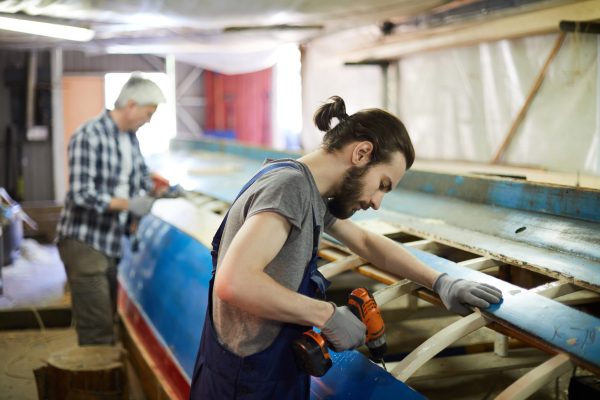 Drilling work Young man with electric drill standing by unfinished structure of vessel and drilling one of its wooden planks
