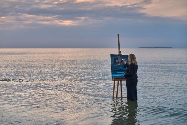 Serene seascape: woman painting in gentle ocean waters at sunset Mature female artist stands in calm ocean waters, painting on easel. Tranquil sunset enhances serene ambiance, blending creativity with nature’s beauty